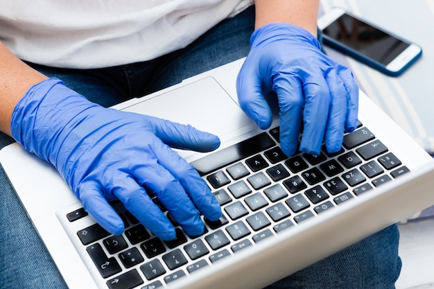 Chiuda sulla donna lavoratrice a casa che scrive sul computer portatile con i guanti chirurgici