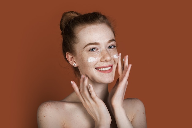 Chiuda sulla foto di una donna lentigginosa allo zenzero con spalle nude che applica crema idratante sul viso sorridente sulla parete rossa dello studio