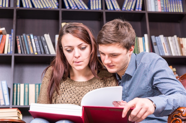 Chiuda sulla giovane coppia dolce che legge un libro alla biblioteca seriamente davanti agli scaffali.