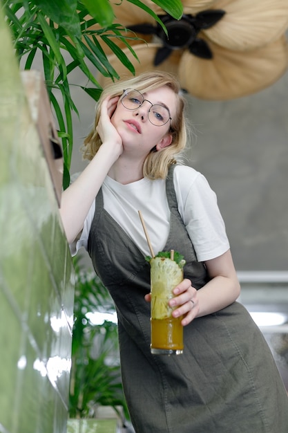 Chiuda sulla giovane donna allegra in vestito che guarda felicemente a porte chiuse con grandi foglie verdi sullo sfondo in un accogliente caffè verde. Ragazza con gli occhiali. tenendo un drink tra le mani