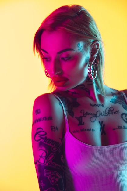 Chiuda sulla giovane donna caucasica in costume da bagno che posa sul fondo giallo dello studio di pendenza alla luce al neon. Bellissimo modello con tatuaggi. Emozioni umane, vendite, concetto di annuncio. Resort e vacanza, estate.