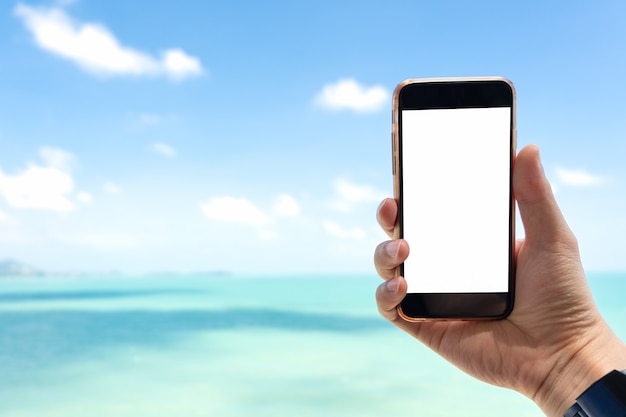 Chiuda sulla mano dell&#39;uomo che tiene smartphone nero sul bello mare blu calmo e sul fondo bianco del cielo.