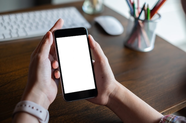 Chiuda sulla mano della donna facendo uso di un telefono cellulare con lo schermo in bianco alla mattina nella casa.