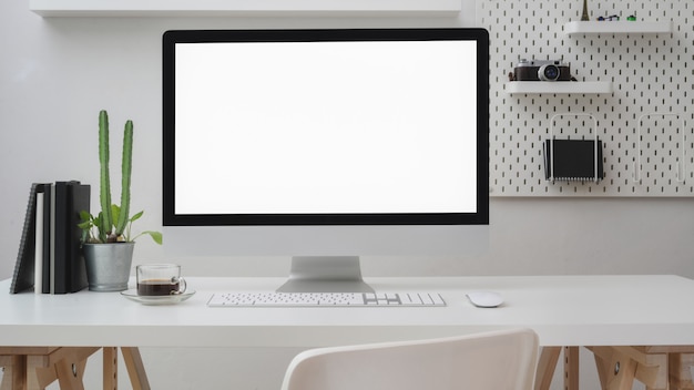 Chiuda sulla vista dell'area di lavoro con il computer dello schermo in bianco, gli articoli per ufficio, la decorazione e lo scaffale sullo scrittorio bianco con la parete bianca