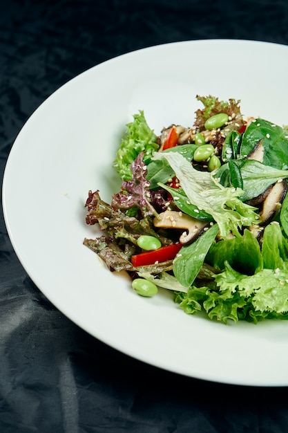 Chiuda sulla vista su insalata con spinaci, funghi, peperone dolce, fagioli e piselli in una ciotola bianca su una tavola nera. Alimenti sani e dietetici. Vegetariano