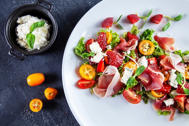 Chiuda sulla vista su insalata fresca e estiva con il pomodoro ciliegia, la fragola, il jamon e il microgreen. Piatti gustosi piatti laici. Cibo per pranzo sano e delizioso. Copia spazio. Vista dall'alto