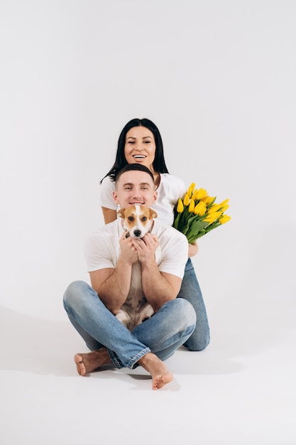 Chiuda sulle giovani coppie del ritratto che si siedono e che abbracciano, tenendo i fiori e il cane gialli in studio su fondo bianco. coppia che si abbraccia con un'espressione amorosa sognante. Famiglia amorevole. Festeggiamo la festa della donna.