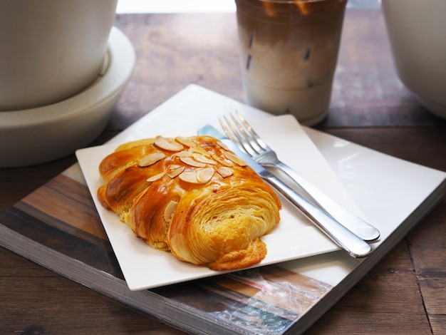 Chiuda sulle mandorle del pane con la forchetta ed il coltello sopra il piatto bianco sul libro e sul vetro del caffè del latte ghiacciato al caffè. Colazione al mattino o pausa caffè.