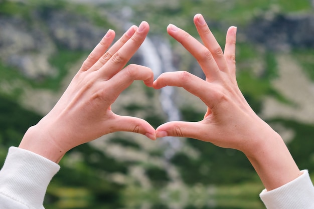 Chiuda sulle mani della donna che mostrano un cuore della cascata e dell'acqua trasparente, pulita, del lago con lo spazio della copia. Vocazione nel concetto di montagna. Solitudine con la natura.