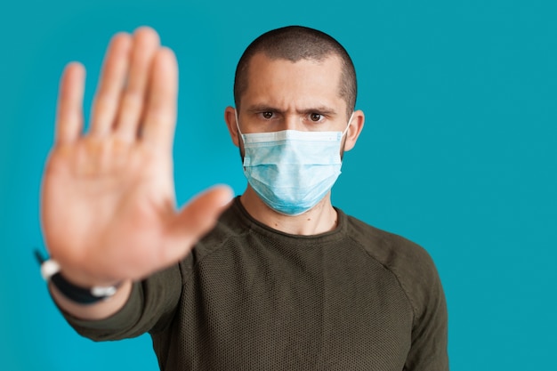 Chiudere la foto di un uomo in maglione che indossa una maschera medica su un muro blu studio gesticolando stop e segno di rifiuto con il palmo
