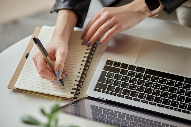 Chiudere la mano della donna utilizzando il computer portatile e prendere appunti nel taccuino seduto sul posto di lavoro a casa wo remoto