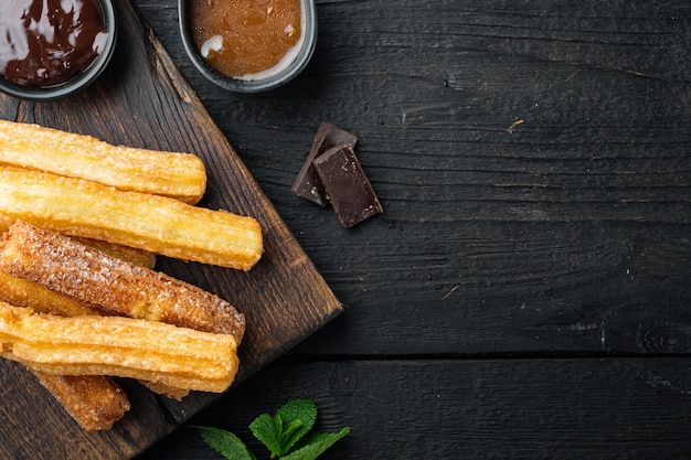 Churros di dessert tradizionali spagnoli con zucchero e cioccolato, sul fondo della tavola in legno nero, vista dall'alto laici piatta con spazio per testo