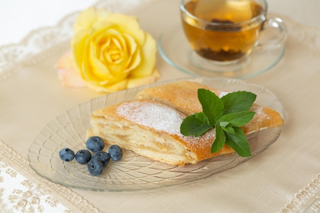 Ci sono due fette di torta di mele, mirtilli e foglie di menta sul piatto una tazza di tè accanto e il tavolo è decorato con una rosa viva