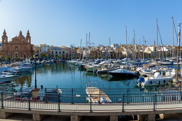 Ci sono molti yacht ormeggiati nel porto sullo sfondo della città maltese con vecchi edifici e palme in una giornata di sole estivo.