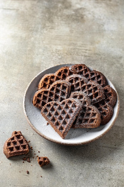 Cialde al cioccolato a forma di cuore cosparse di zucchero a velo nel piatto su fondo strutturato. Concetto per San Valentino