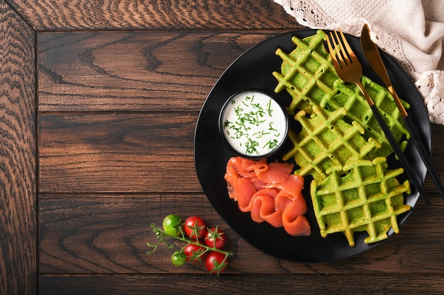 Cialde belghe verdi Spinaci o aglio selvatico o cialde al pesto con salmone rosso e salsa di panna su piatto nero su sfondo di legno rustico scuro Deliziosa colazione spuntino brunch Vista dall'alto Mock up