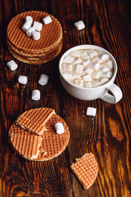 Cialde olandesi fatte in casa con uno rotto con tazza bianca di cacao con marshmallow e pila di cialde. Orientamento verticale.