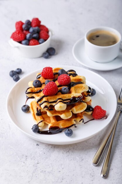 Cialde viennesi o belghe con frutti di bosco freschi lamponi e mirtilli e salsa di cioccolato su un piatto bianco e una tazza di caffè Dessert tradizionale Primo piano sfondo bianco