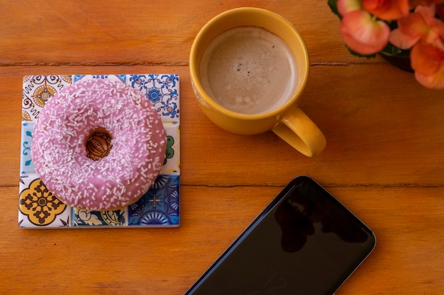 Ciambella glassata rosa sul tavolo di legno, tazza di caffè e cellulare - pausa o colazione