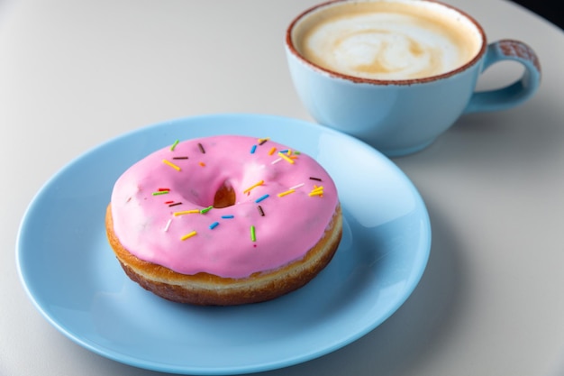 Ciambella ricoperta di glassa rosa e granelli multicolori su piatto blu e tazza di cappuccino