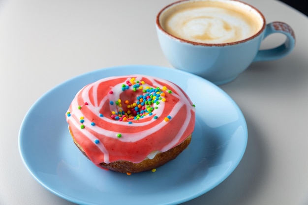 Ciambella ricoperta di glassa rossa e granelli multicolori su piatto blu e tazza di cappuccino