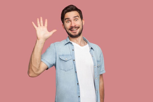 Ciao piacere di conoscerti. Ritratto di sorpreso bel giovane barbuto in camicia stile blu in piedi e guardando la telecamera con sorriso a trentadue denti e saluto. foto in studio al coperto, isolata su sfondo rosa