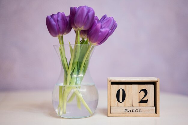 Ciao primavera Calendario cubico in legno con data del 2 marzo circondato da bouquet di tulipani viola con foglie verdi in vaso di vetro