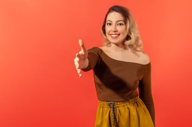 Ciao Ritratto di donna con capelli biondi in abiti casual che danno la mano alla stretta di mano e sorridente amichevole collaborazione guardando la telecamera Studio indoor girato isolato su sfondo rosso