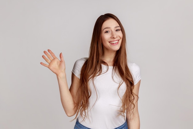 Ciao. Ritratto di donna felice in piedi con la mano alzata che dice ciao alla fotocamera, salutando gli ospiti con un sorriso amichevole, indossando una maglietta bianca. Studio indoor girato isolato su sfondo grigio.