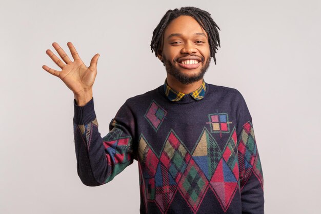 Ciao tu. Uomo africano amichevole positivo con i dreadlocks e la barba che agita la mano che ti saluta con un sorriso a trentadue denti sul viso, bello incontrarsi, uscire. Studio indoor girato isolato su sfondo grigio