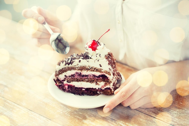 Cibo, cibo spazzatura, cucina, cottura al forno e concetto di vacanze - primo piano di donna che mangia una torta di ciliegie al cioccolato con un cucchiaio e si siede al tavolo di legno su uno sfondo di luci natalizie