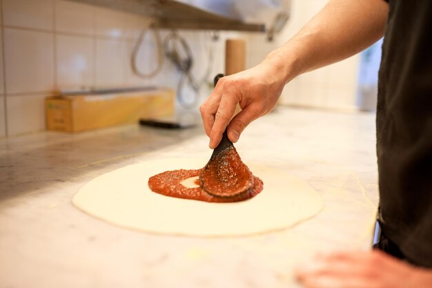 cibo, cucina culinaria, cucina italiana, persone e concetto di cucina - cucina con un cucchiaio applicando la salsa di pomodoro all'impasto della pizza cruda in pizzeria