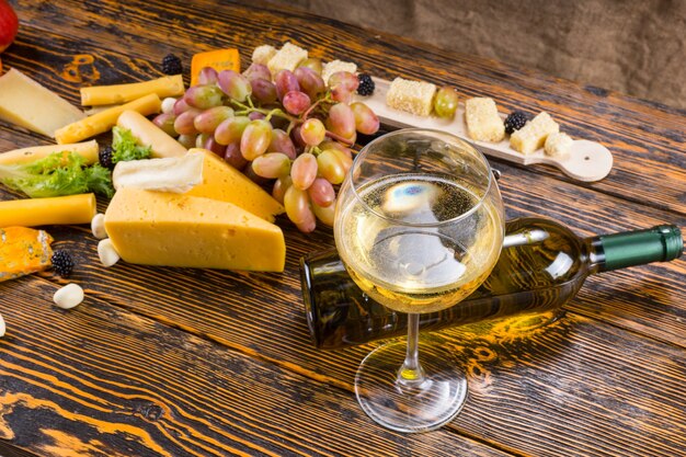 Cibo Gourmet Still Life - Close Up di bicchiere di vino bianco con bottiglia caduta tra varietà di formaggi e frutta fresca su tavola in legno rustico con spazio di copia