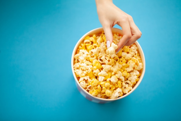 Cibo. Mano della donna che prende popcorn dal secchio. Secchio di popcorn su blu. Cinema