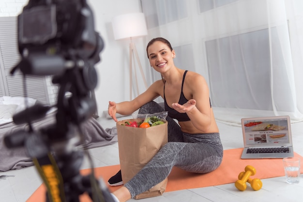 Cibo salutare. Bella giovane donna atletica esuberante che sorride e che mangia un pacchetto con le verdure mentre si siede sul tappeto e fa un video per il suo blog