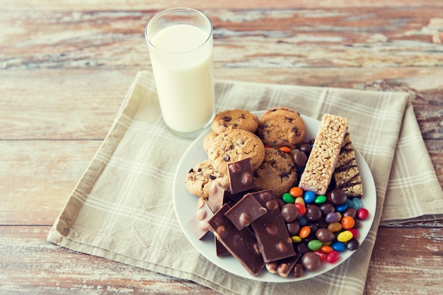 cibo spazzatura, dolci e concetto di alimentazione malsana - primo piano di caramelle, cioccolato, muesli e biscotti con bicchiere di latte sul piatto