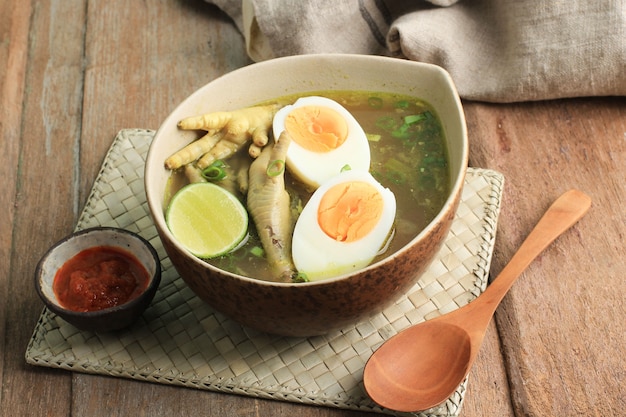 Cibo tradizionale indonesiano chiamato Soto Ceker Ayam (zuppa di pollo con coscia di pollo) su ciotola di ceramica marrone, con Sambal, uova sode e lime