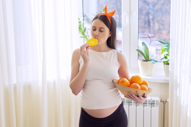 Cibo vitaminico sano, bella giovane donna incinta felice con arance a casa vicino alla finestra. Gravidanza, vitamine, concetto di maternità