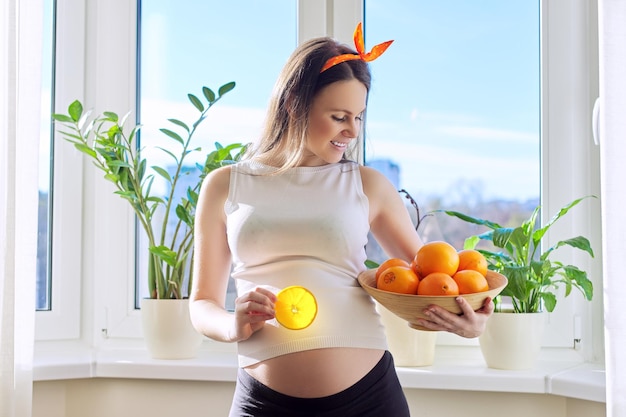 Cibo vitaminico sano, bella giovane donna incinta felice con arance a casa vicino alla finestra. Gravidanza, vitamine, concetto di maternità