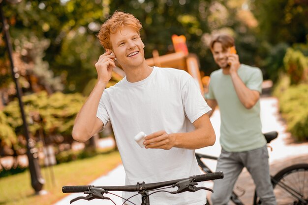 Ciclista allegro che si mette un auricolare wireless nell'orecchio mentre il suo amico chiacchiera sullo smartphone