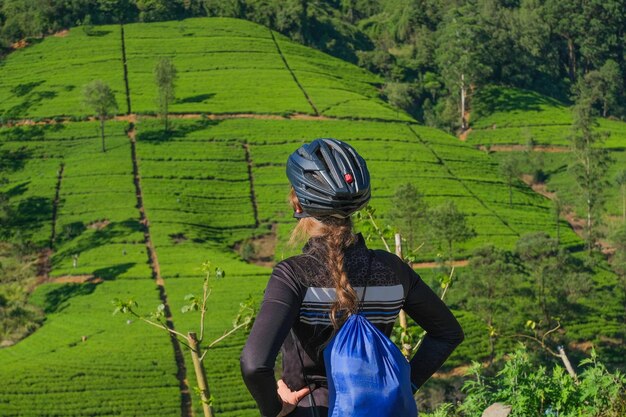 Ciclista femminile dei capelli biondi in casco che esamina le piantagioni di tè e la vista posteriore della foresta