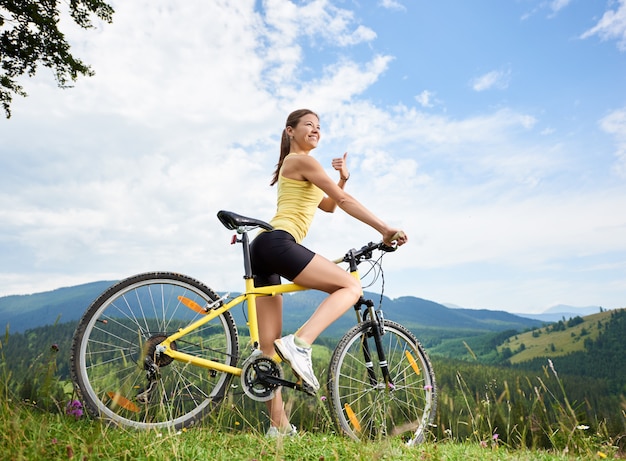 Ciclista femminile felice attraente che guida su mountain bike giallo su una collina erbosa, mostrando i pollici, godendo la giornata estiva in montagna. Attività sportiva all'aperto, concetto di lifestyle