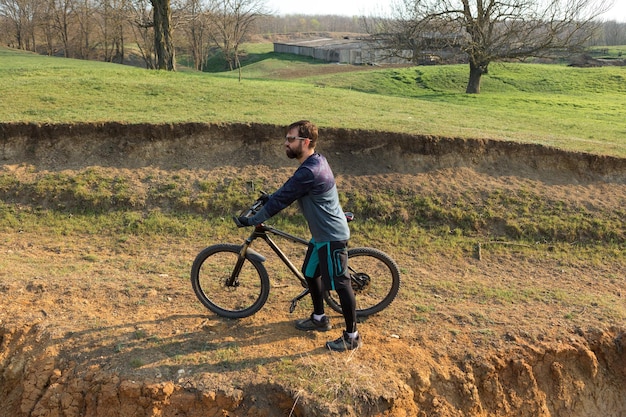 Ciclista in pantaloncini e maglia su una moderna bici hardtail in carbonio con una forcella a sospensione pneumatica in piedi su una scogliera sullo sfondo di una fresca foresta verde primaverile