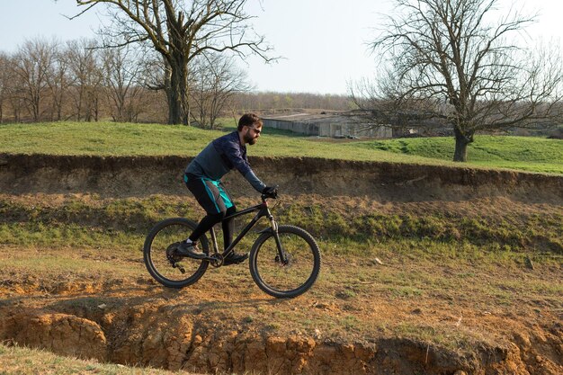 Ciclista in pantaloncini e maglia su una moderna bici hardtail in carbonio con una forcella a sospensione pneumatica in piedi su una scogliera sullo sfondo di una fresca foresta verde primaverile