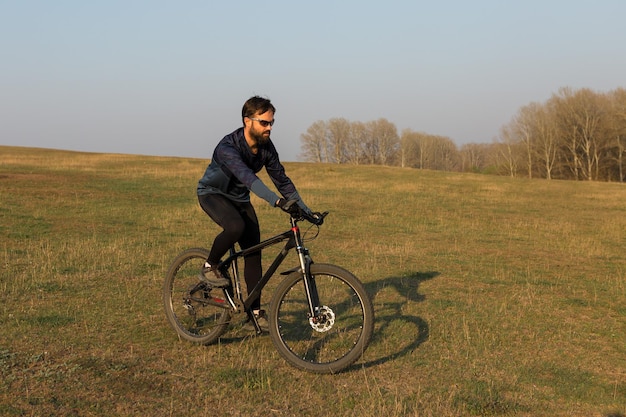 Ciclista in pantaloncini e maglia su una moderna bici hardtail in carbonio con una forcella a sospensione pneumatica in piedi su una scogliera sullo sfondo di una fresca foresta verde primaverile