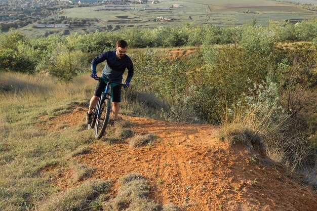 Ciclista in pantaloncini e maglia su una moderna bici hardtail in carbonio con una forcella a sospensione pneumatica in piedi su una scogliera sullo sfondo di una fresca foresta verde primaverile