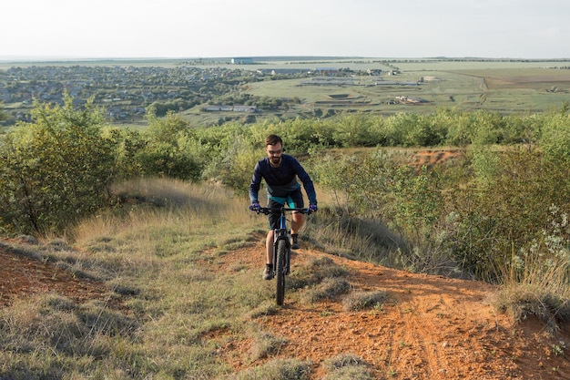 Ciclista in pantaloncini e maglia su una moderna bici hardtail in carbonio con una forcella a sospensione pneumatica in piedi su una scogliera sullo sfondo di una fresca foresta verde primaverile