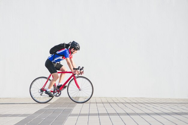 Ciclista in un casco, abiti da ciclismo atletici e uno zaino cavalca su un muro bianco