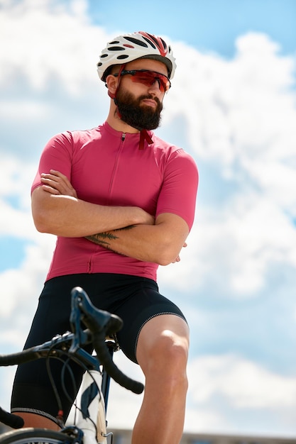 Ciclista maschio forte con occhiali sportivi e casco protettivo che cammina con la sua bici nella foresta per fare una pausa dopo aver guidato il cielo blu sullo sfondo