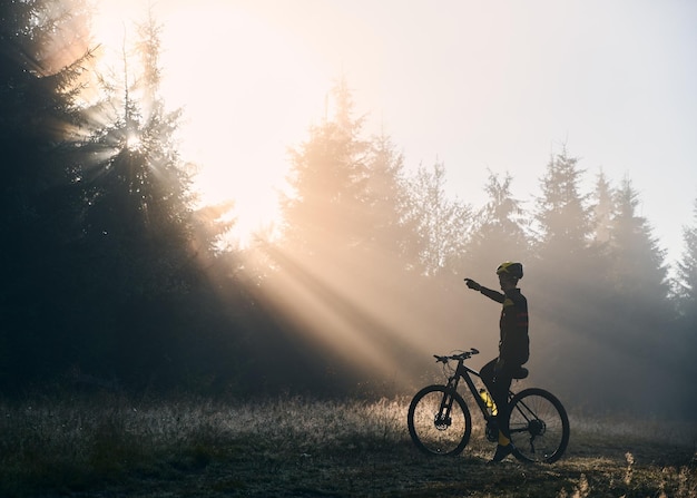 Ciclista maschio seduto in bicicletta e guardando l'alba nella foresta